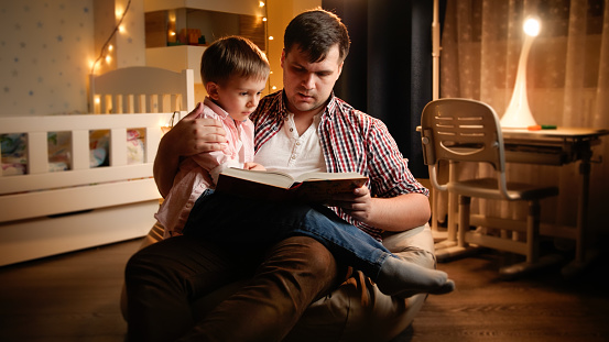 Young father sitting in bedroom with his little son and reading fairy tale book. Concept of child education and family having time together at night.