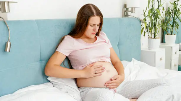 Young pregnant woman feeling unwell and abdominal pain sitting on bed. Healthcare and intoxication during pregnancy.