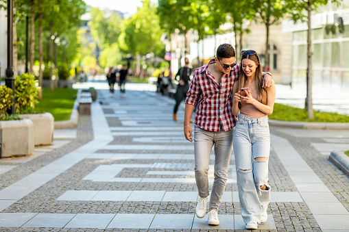 Young couple using smartphone outdoors. Joyful smiling woman and man looking at mobile phone in a city. Love, technology, communication, connection, friends, students, summer travel concept