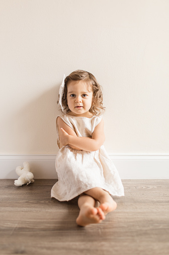 Two-Year-Old Toddler Girl Sitting on the Floor in a Cream-Colored Linen Dress & a Bow at Home During COVID-19