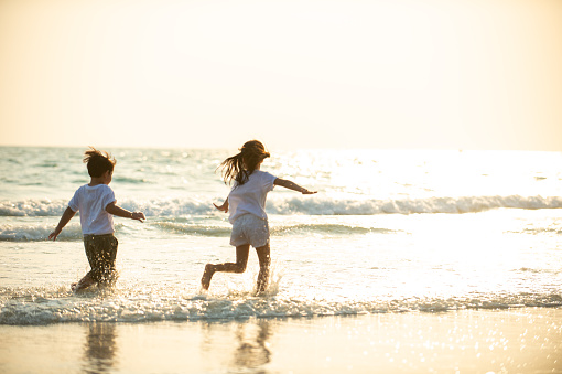 Happy Asian family on beach travel. Little brother and sister running and playing together on the beach at summer sunset. Adorable child girl and boy sibling enjoy and having fun in outdoor summer holiday vacation