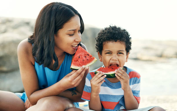 schuss einer mutter und sitzen und genießen einige wassermelone am strand - fruit flavor stock-fotos und bilder