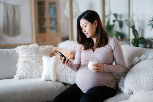 美麗的亞洲年輕孕婦在舒適的家客廳的沙發上放鬆。喝一杯鮮奶，用智能手機。懷孕期間的健康、健康飲食生活方式 - healthy diet of a pregnant woman 個照片及圖片檔