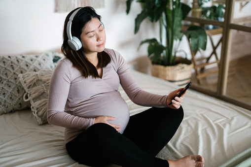 Young Asian pregnant woman with her eyes closed cradling her belly, relaxing on bed at cozy home. Holding smartphone, listening to calming music on headphones. Encourages the stimulation of brain development of her unborn baby. Healthy pregnancy lifestyle