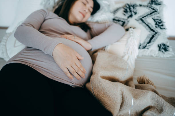 Asian pregnant woman holding her belly, sleeping on bed. Comfortable sleeping positions during pregnancy. Insomnia, sleeping disorder and physical discomfort during pregnancy