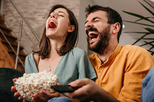 Beautiful and cheerful young adult couple enjoying their time, laughing while being squeezed together on the sofa. Both eating popcorns and watching comedy.