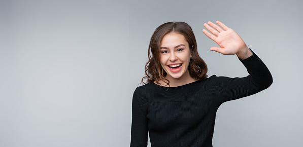 Hi what is up. Portrait of friendly young brunette woman smiling and saluting, waving hand to say hello, greeting friends, say hi and welcome, goodbye gesture, standing against gray background