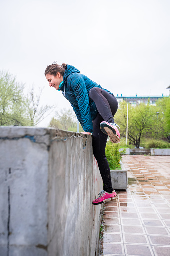 Girl wearing jacket on a wet day, going over a concrete wall in residential area, leaning with both hands and lifting her leg