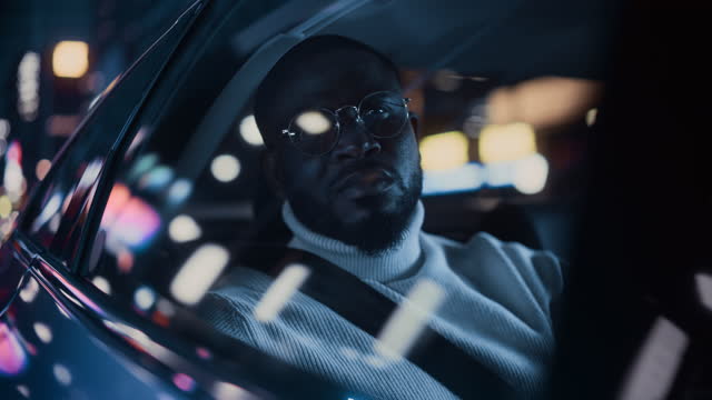 Stylish Black Man in Glasses is Commuting Home in a Backseat of a Taxi at Night. Handsome Male Passenger Looking Out of Window while in a Car in Urban City Street with Working Neon Signs.