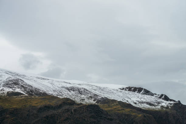 istock Minimalist highlands landscape with snow-capped mountains under cloudy sky. Snowbound high mountain range in overcast weather. Atmospheric minimalism with green mountains, white snow and gray sky. 1319003677