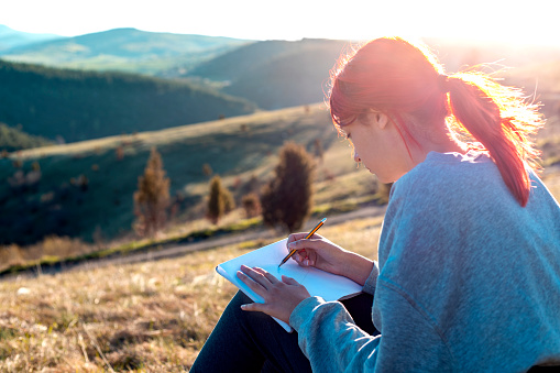 Young woman hiker enjoy the view at sunset seaside mountain peak. She siting on a meadow and sketching a sunset.