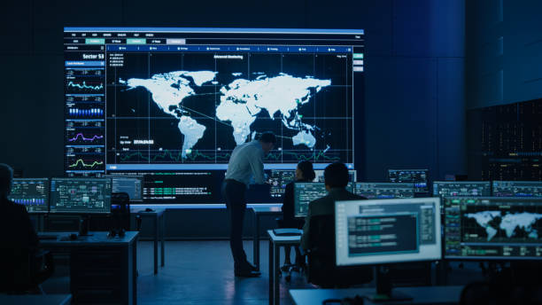 istock Project Manager Uses Digital Tablet Talks with Colleague. Big Screen Display Showing Infographics and Global World Map Data. Telecommunications Control Monitoring Room with People Working 1319010432