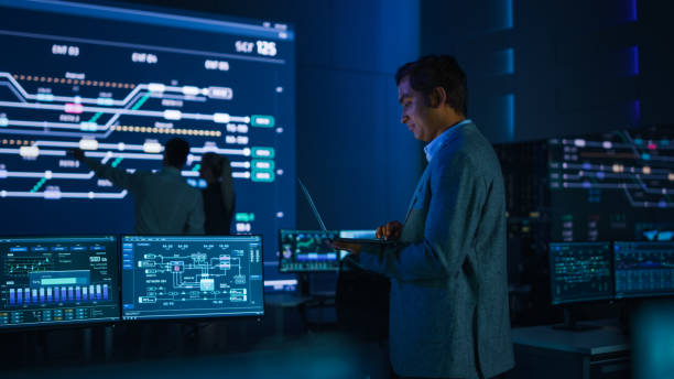istock Handsome Confident Male Project Manager works on a Laptop Standing in System Control Room. In Background Big Screen Showing Infographics of Infrastructure, Charts, System Analysis. 1319010472