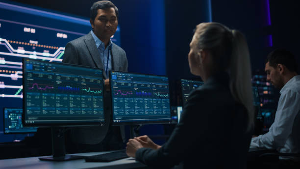 istock Confident Female Specialist Works on Computer Talks with Team Leader in Big Infrastructure Control Room. Business Woman Professional Uses Computer Showing Charts, Information. Monitoring Room Teamwork 1319010501