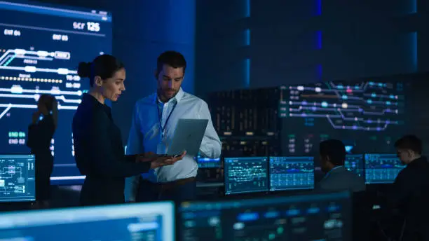 Photo of Project Leader and Chief Executive Discuss Data Shown on a Laptop. In Background Big Screen Showing Infographics of Infrastructure, Charts, System Analysis. Telecommunications Control Room