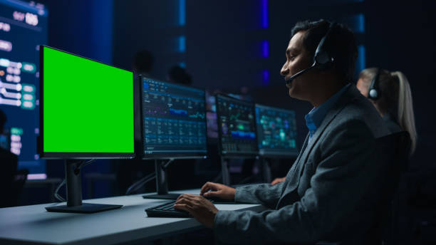 istock Customer Support Specialist Talks to Client on Headset Works on Green Chroma Key Screen Computer in Big Infrastructure Control Room. Specialists Use Computers Showing Graphs. Talk to Investors 1319010519