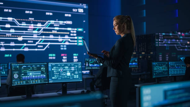 Beautiful Confident Female Project Manager works on a Laptop Standing in Telecommunications System Control Room. In Background Screen Showing Infographics of Infrastructure, Charts, System Analysis beautiful confident female project manager trabaja en una computadora portátil de pie en la sala de control del sistema de telecomunicaciones. en la pantalla de fondo que muestra infografías de infraestructura, gráficos, análisis del sistema