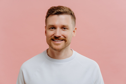 Portrait of handsome pleasant unshaven redhead man expressing positiveness while looking at camera with smile, happy guy in white tshirt feeling joyful while posing against pink studio background.