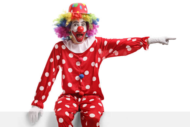 istock Funny clown sitting on a white panel and pointing to the side 1319016777