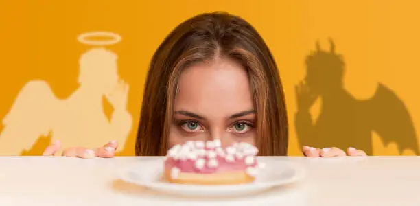 Hungry woman looking at yummy donut from under table, angel and devil tempting her, orange studio background, collage. Panorama. Concept of choice between healthy diet and unhealthy foods