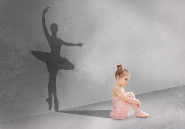 Cute little girl sitting on floor in dance studio, shadow of ballet dancer on grey wall, dreaming to become dancing star Cute little girl sitting on floor in dance studio, shadow of ballet dancer on grey wall, dreaming to become dancing star in future, imagining her future success and fame, creative collage ballerina shadow stock pictures, royalty-free photos & images