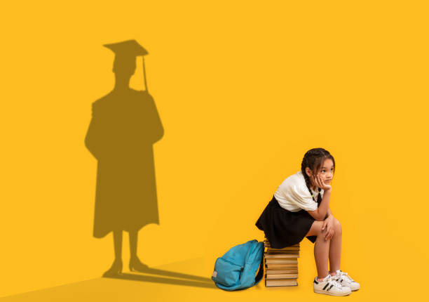 Pensive Asian schoolgirl sitting on stack of books, dreaming of college graduation, shadow of university student on wall Thoughtful Asian schoolgirl sitting on stack of books, dreaming of college graduation, shadow of university student in mortarboard and mantle on orange studio wall. Success and potential concept junior level stock pictures, royalty-free photos & images