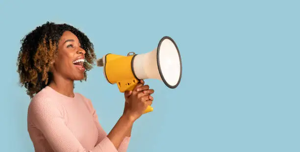 Announcement Concept. Cheerful Black Woman Shouting With Megaphone In Hands, Curly African American Lady Holding Loudspeaker, Sharing News, Standing Isolated On Blue Background, Panorama, Copy space