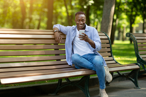 Smiling black man sitting on bench at park and using his smartphone, copy space