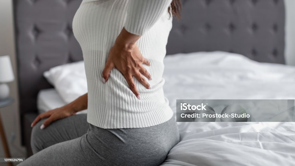 Woman with side back pain sitting on bed at home People and Health Problem Concept. Coseup of sick young black woman suffering from acute pain in left side, sitting on bed at home, touching lower back with hand, free copy space Chronic Illness Stock Photo