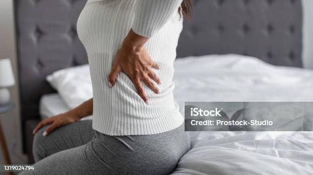 Woman With Side Back Pain Sitting On Bed At Home Stock Photo - Download Image Now - Chronic Illness, Backache, Women