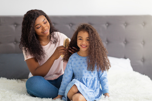 Loving black mom brushing her pretty daughter's hair on bed at home. Cute little child and caring mother doing hairstyles, having fun together, playing beauty salon, spending spa day indoors