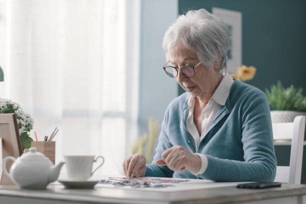 senior woman sitting at desk and solving a puzzle - jigsaw puzzle solution one person people imagens e fotografias de stock