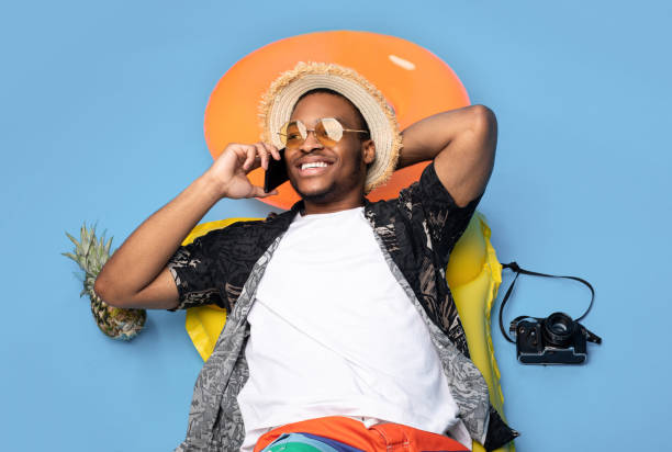 Funky black guy in trendy summer clothes lying on inflatable lilo, speaking on smartphone over blue background, top view