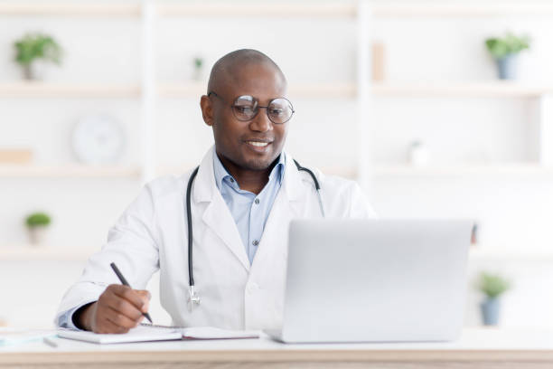 istock Professional help concept. Friendly black male doctor in white unifrom sitting at table with laptop at his office 1319026123