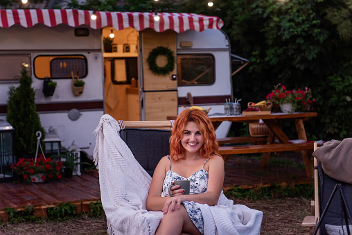 Young red-haired woman sits on a wooden deck chair outside a trailer home. Girl holds in her hands a gray cup of cocoa, tea, wrapped in a white warm blanket, enjoys outdoor recreation outside the city