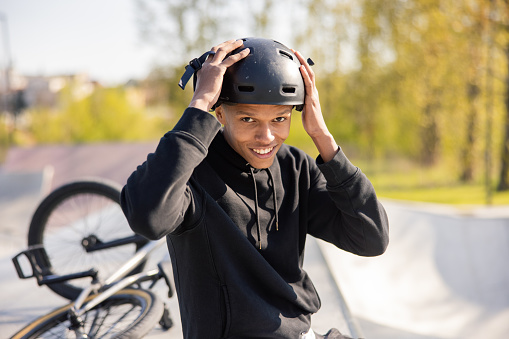 A young boy sits on a ramp at a skatepark with his bike lying wheel up behind him. The guy has finished riding and takes his black helmet off his head and slides it off with two hands