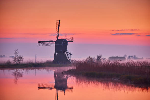 Windmill The Broekmolen during sunrise on an early morning. The Broekmolen in the Dutch village of Streefkerk, municipality of Molenlanden, is a seesaw mill with open shovel wheel and was built in 1581 to grind the Kortenbroek department of the Streefkerk polder. The mill was in operation until 1951. In 1957 the Kleine Molen was decommissioned, but the polder was obliged to keep the Broekmolen grinding. The sky is beautifully colored in orange, pink and purple. In the background is a big forest and there is a layer of fog over the grass. The windmill is standing next to the water that reflects its surroundings.