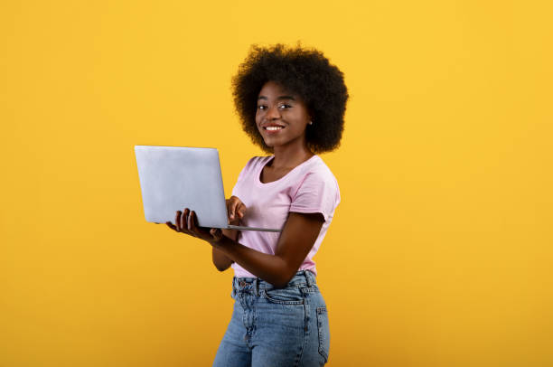istock Online work. Excited black woman holding and using newest laptop computer, standing over yellow studio background 1319029705