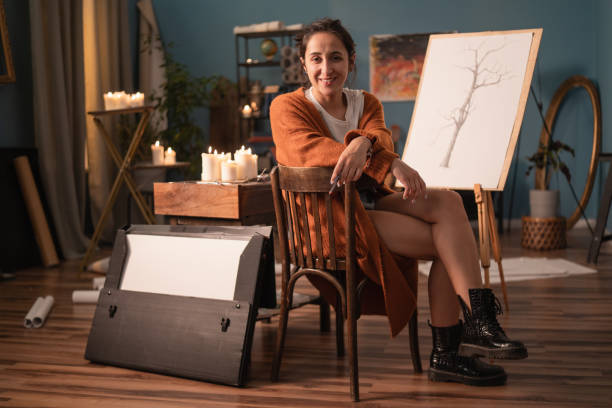a talented artist smiles passionately as she sits in her art studio on a chair with a folder of drawing blocks, pencils, and a new sketch in the background - drawing sketch artist charcoal drawing imagens e fotografias de stock