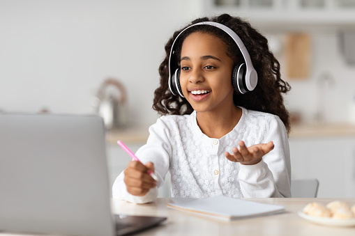 Joyful african american girl having online lesson from home, using wireless headphones and laptop, kitchen interior. Cute black schooler having video chat with teacher, copy space