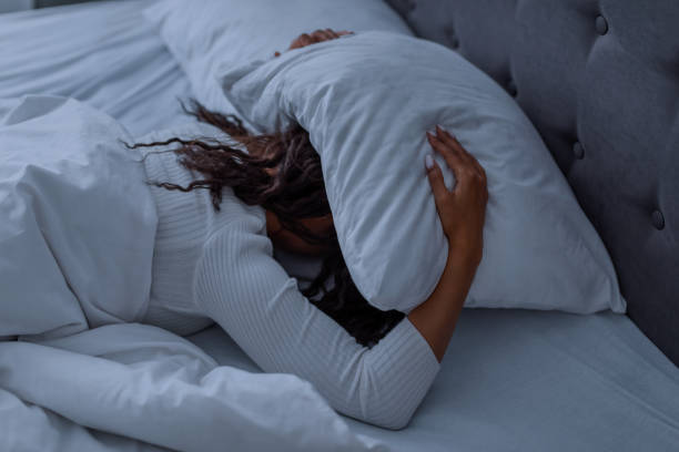 stressed black woman covering head under pillow lying in bed - female emotional stress african ethnicity loss imagens e fotografias de stock