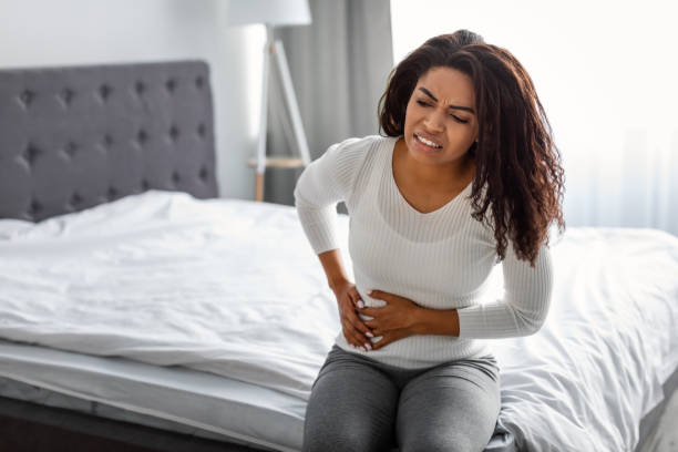 Woman with side pain sitting on bed at home People, Healthcare and Health Problem Concept. Portrait of upset sick young black woman suffering from acute pain in right side, sitting on bed at home, free copy space. Abdomen liver pain cirrhosis stock pictures, royalty-free photos & images