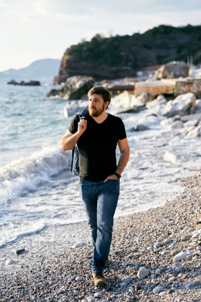 istock Pensive man walking on a pebble beach with a denim jacket over his shoulder against the background of the sea and rocks 1319038825