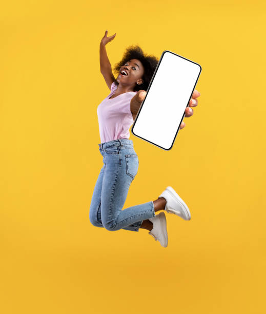istock Overjoyed african american young lady showing empty smartphone screen while jumping up over yellow studio background 1319039339