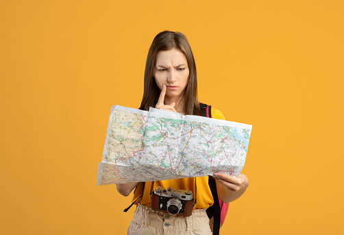 Confused woman traveler with camera looking at city map, got lost, yellow studio background, copy space. Upset young lady backpacker lost in city, checking map. Navigation, trekking during travelling