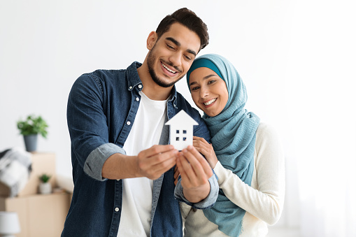 Young loving muslim couple middle eastern man and woman in hijab showing paper house at camera and smiling, real estate and relocation concept, copy space. Muslim family moving