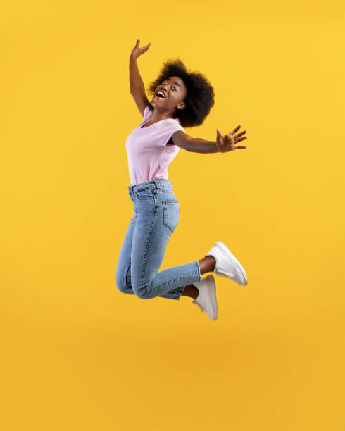 positivamente afroamericana tupida dama saltando y posando en el aire, divirtiéndose sobre el fondo amarillo del estudio, cultivo - saltar actividad física fotos fotografías e imágenes de stock