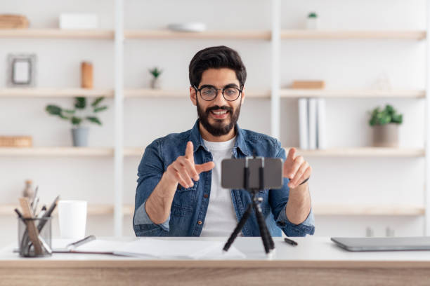 istock Positive arab male influencer broadcasting from home, using smartphone and tripod, sitting at table in living room 1319041832