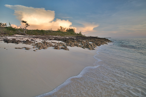 Morning sun lighting up clouds over a deserted sand and rock beach in the Bahamas.