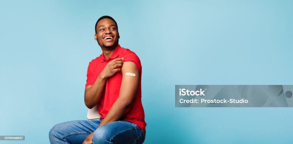 African American Guy Showing Arm After Vaccine Injection, Blue Background I'm Vaccinated. Joyful African American Guy Rolling Up Sleeve Showing Arm After Vaccine Injection Shot With Plaster On Blue Background. Health, Immunization And Vaccination. Panorama, Empty Space Vaccination Stock Photo